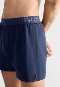 Boxers bleu marine en tissu doux avec une taille élastique ornée de "SLG" brodé. Comprend des fentes sur les côtés pour plus de flexibilité.