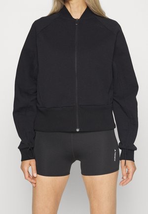 Femme portant une veste noire zippée et un short de sport noir ajusté avec le logo "TECHFIT" sur la cuisse gauche, debout devant un fond blanc.