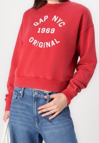 Sudadera roja con el texto blanco "GAP NYC 1969 ORIGINAL", cuello redondo, puños acanalados, combinada con vaqueros de mezclilla azules. Tejido suave y casual.