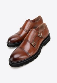 Bruine leren schoenen met een gepolijste afwerking, voorzien van twee zilveren gespen, een ronde neus en een dikke zwarte rubberen zool.