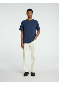 Marinblå t-shirt med kort ärm i bomull, i kombination med krämfärgade byxor med vida ben och svarta skor, stående mot en enfärgad bakgrund.