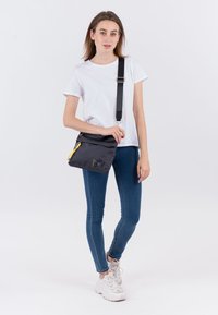 Borsa a tracolla nera realizzata in tessuto resistente, con dettagli in giallo della zip, chiusura a patta e spallaccio regolabile.