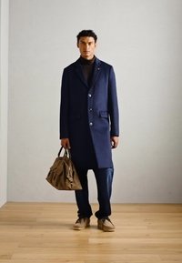 Abrigo largo de color marino con solapas, tres botones y bolsillos de parche, combinado con un suéter de cuello alto oscuro, pantalones de mezclilla y un bolso marrón.