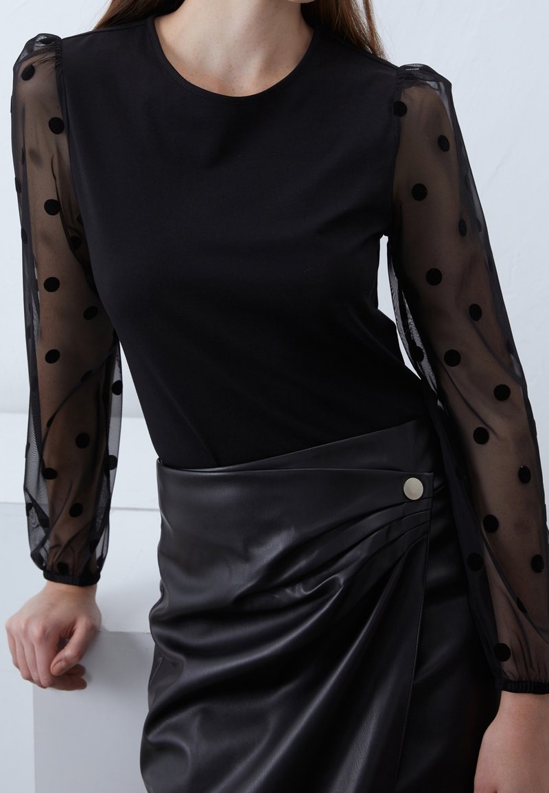 Top nero con maniche a sbuffo trasparenti decorate con pois neri, abbinato a una gonna a vita alta in pelle nera con tessuto drappeggiato e dettaglio con bottone.