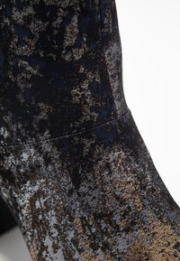 Bottine noire avec un tissu texturé comportant des motifs mouchetés en bleu, or et argent. Talon épais, silhouette élégante.