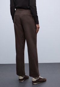 Pantalons marron à jambes larges avec une texture lisse, dotés de deux poches arrière et d'une taille haute. Assortis à des chaussures sombres.