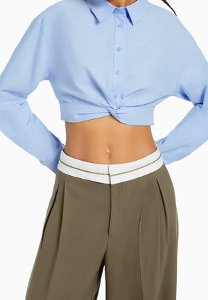Kobieta ubrana w błękitną, krótką koszulę zapinaną na guziki z frontowym wiązaniem oraz oliwkowozielone spodnie z wysokim stanem i białym paskiem w talii.
