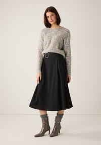 Grauer Pullover mit Leopardenmuster, kombiniert mit einem schwarzen A-Linien-Rock, der einen breiten Gürtel und seitliche Taschen aufweist. Das Model trägt graue gerippte Socken und graue Stiefeletten mit Absatz.