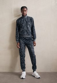 Mørkegrå treningstøy med leopardmønster, bestående av en glidel�åsjakke og elastiske bukser, kombinert med hvite sportssko.
