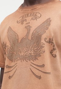 Smėlio spalvos marškinėliai su dideliu tekstūruotu erelio grafiniu piešiniu centre, virš kurio yra žodis „DIESEL“ ir metai „1978“.