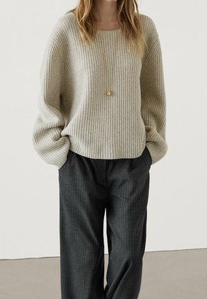 Person wearing a beige ribbed sweater, black pinstripe pants, and a gold pendant necklace, standing with hands in pockets.