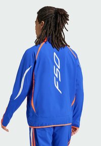 Chaqueta deportiva azul con acentos naranjas y detalles en blanco. Presenta un gran logo "F50" en la parte posterior. El material parece suave y ligero.