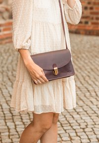 Frau in einem cremefarbenen, strukturierten Kleid, die eine dunkelbraune Leder-Umhängetasche mit goldener Schnalle hält, steht auf Kopfsteinpflaster in der Nähe einer Ziegelmauer.