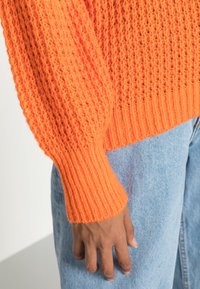 Gebreide oranje trui met een gestructureerd ontwerp en geribde manchetten, gecombineerd met lichtblauwe denimjeans, die een casual outfitdetail toont.