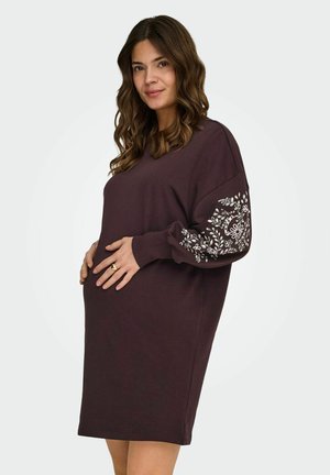 Gravid kvinde i en løs, mørkelilla kjole med hvid blomstret broderi på ærmerne, blidt holdende om sin mave og smilende blidt.