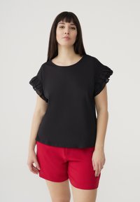 Maglietta nera a maniche corte con dettagli arricciati; abbinata a pantaloni corti rossi. Tessuto liscio; scollatura rotonda con vestibilità rilassata.