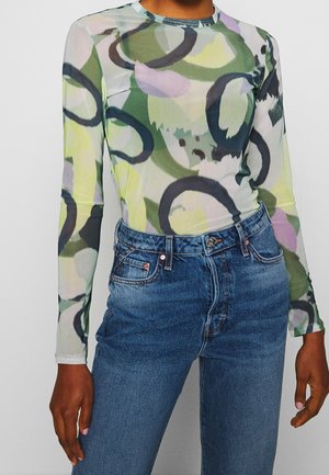 Nainen pukeutuneena vartalonmyötäiseen, pitkähihainen abstraktikuosiseen yläosaan, jossa on vihreitä, mustia ja purppuran sävyjä, yhdistettynä korkeavyötäröisiin sinisiin farkkuihin.