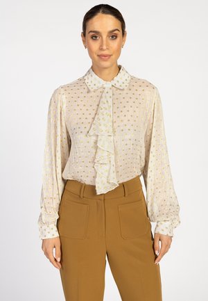 Crème blouse met een goudkleurig stippenpatroon en ruches aan de voorkant, gecombineerd met hoge, camelkleurige broeken met twee zakken.