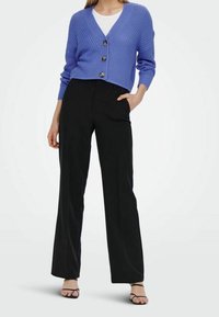 Mujer vistiendo un cárdigan de punto azul con botones, sobre una blusa blanca, pantalones de pierna ancha negros y sandalias negras con tiras, de pie con una mano en el bolsillo.