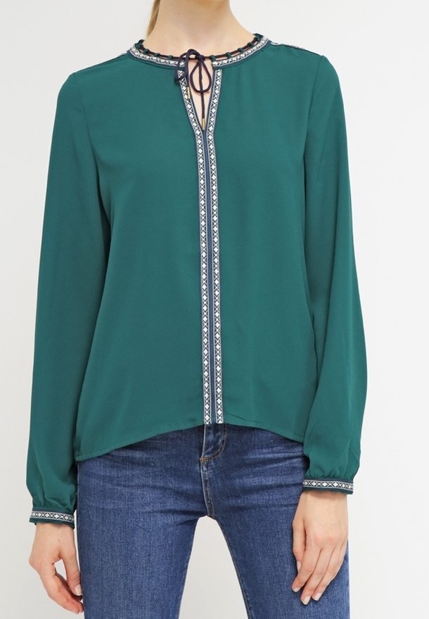 Tealfärgad blus med långa ärmar, rund halsringning, framtida knytning och kontrasterande broderad trim längs knäppningen och ärmsluten. Mjuk, lättviktig tyg.