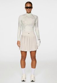 Långärmad, ljusgrön topp med abstrakt mönster, kombinerad med en plisserad beige kjol; vita strumpor och skor, bär solglasögon och håller i en handske.