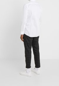 Camisa blanca de manga larga con cuello abotonado, combinada con pantalones negros a cuadros y zapatillas blancas. Enfoque en un ajuste entallado y líneas limpias.