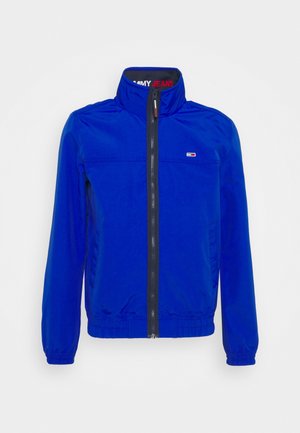 Chaqueta cortavientos azul con un alto cuello, cierre de cremallera, puños elásticos y un parche con logo en el pecho. Tejido suave y ligero.