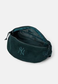 Sac banane en velours côtelé vert foncé avec fermeture éclair ouverte et logo brodé à l'avant, montrant la doublure intérieure vide.