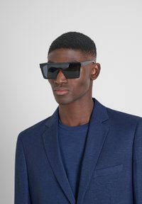 Gafas de sol negras de gran tamaño con forma angular, montura gruesa y lentes oscuros, llevadas con un blazer azul marino y una camisa a juego.