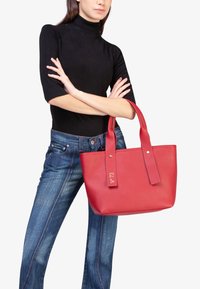 Rode leren tote bag met twee lange handvatten, voorzien van een goudkleurige logo label en een gladde textuur. Draagt met donkere jeans en een zwart top.