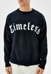 Persona vestida con un suéter negro que tiene el texto blanco "Timeless" en el pecho y agujeros desgastados a lo largo del dobladillo, combinado con pantalones claros.