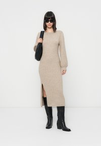 Robe en tricot beige à manches longues et fente latérale, associée à des bottes noires jusqu'au genou et un sac à épaule noir. Texture lisse et design ajusté.