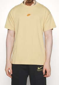 En person iklädd en beige Nike-t-shirt och svarta Nike-shorts med gula logotyper, som står mot en ljus bakgrund.