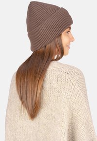 Braune, gerippte Strickmütze mit umgeschlagenem Bündchen, getragen von einem Modell mit glattem Haar. Weiche Textur, eingerahmt von einem hellbeigen Strickpullover.