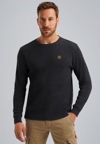 Dunkelgraues Langarmshirt aus strukturiertem Stoff, mit rundem Ausschnitt und einem kleinen gelben Logo-Patch auf der Brust.