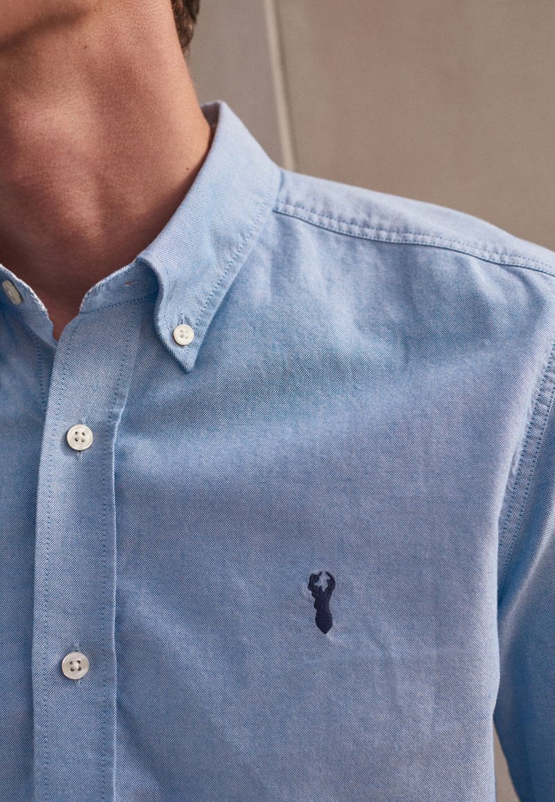 Hellblaues Button-up-Hemd aus Baumwollstoff, mit einem dezenten navyblauen gestickten Logo auf der Brust und weißen Kunststoffknöpfen.