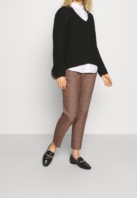 Zwarte gebreide trui met V-hals, witte blouse eronder, bruine geruite broek en zwarte instappers met gouden hardwareaccenten.