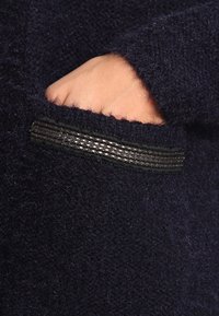 Námořnická tkanina se strukturou a měkkým povrchem, obsahující kapsu s kovovým pruhovaným akcentem. Detail ukazuje ruku částečně uvnitř kapsy.