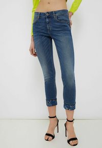 Mujer vestida con jeans azules cortos con puños bordados en flores y sandalias de tacón negro, de pie contra un fondo liso.