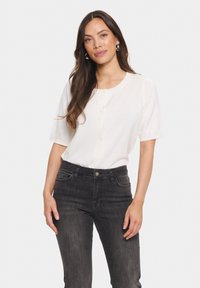Vit kortärmad cardigan med knappar, tillverkad av ett mjukt material. Matchad med mörkgrå tajta jeans, subtila fickor synliga.