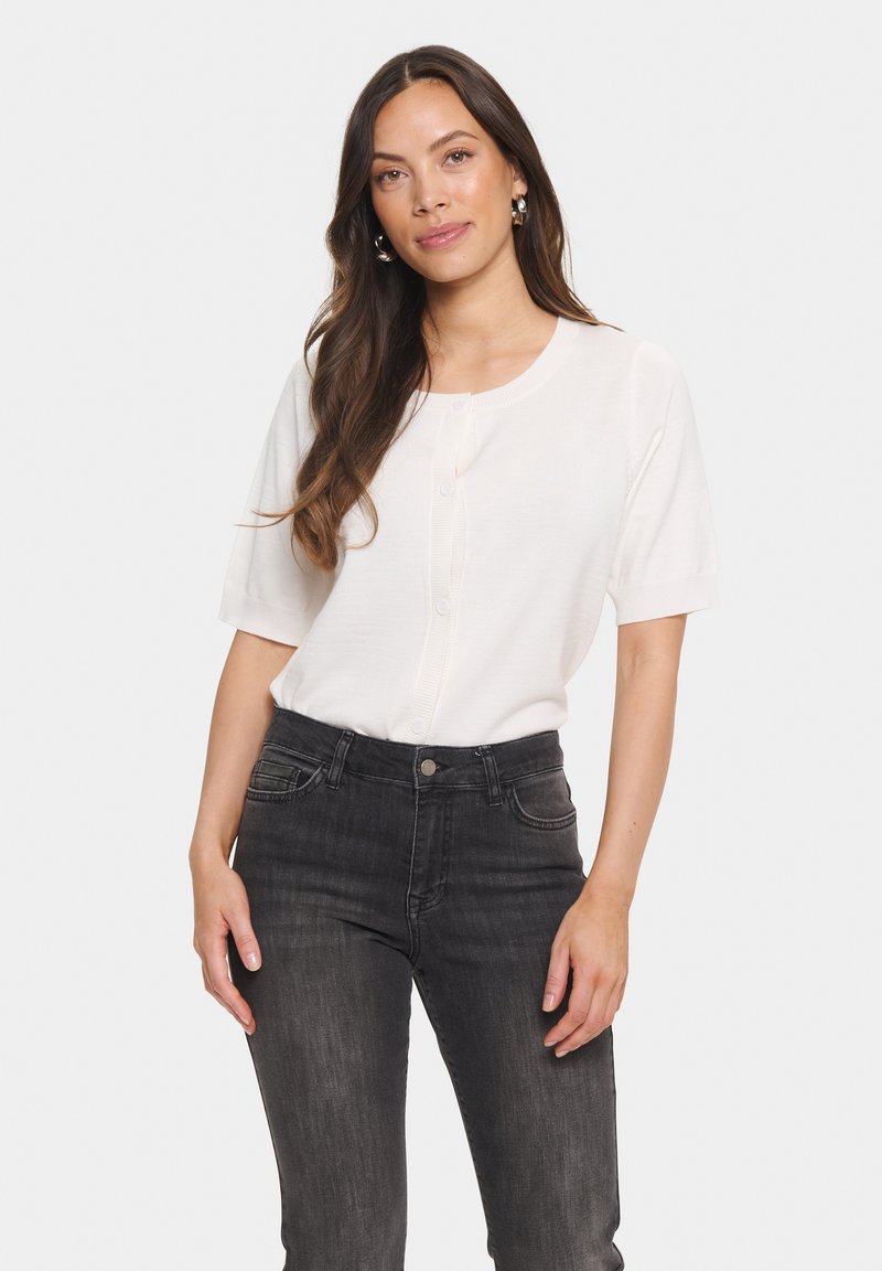 Vit kortärmad cardigan med knappar, tillverkad av ett mjukt material. Matchad med mörkgrå tajta jeans, subtila fickor synliga.