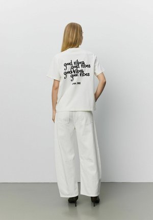 Personne aux longs cheveux blonds portant un t-shirt blanc avec le texte "good vibes" répété et un pantalon blanc ample, debout devant un fond uni.