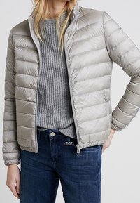 Helle graue Daunenjacke, gestepptes Design, mit Reißverschluss auf der Vorderseite, hoher Kragen, getragen über einem grauen Rippenpullover, kombiniert mit dunkelblauen Jeans.