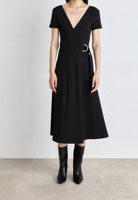 Kobieta w czarnej midi sukience nawiązującej do stylu wrap z krótkimi rękawami i dekoltem w serek, w połączeniu z czarnymi, wskazującymi butami do połowy łydki, stoi na gładkiej podłodze.