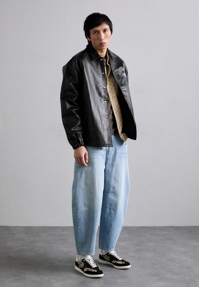 Svart skinnjakke med knappelukking, lys beige mønstret skjorte, vide lyseblå jeans og svarte joggesko med hvite detaljer.