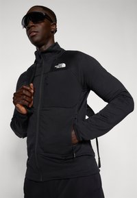 Svart zip-up jakke med strukturert stoff, en brystlomme med glidelås, og North Face-logoen på øvre venstre side.