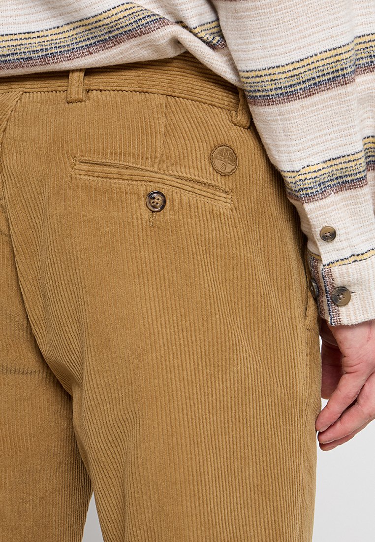 Pantaloni in corduroy marrone con texture a coste verticali, dotati di una tasca posteriore con bottone e un accento di marca sopra la tasca.