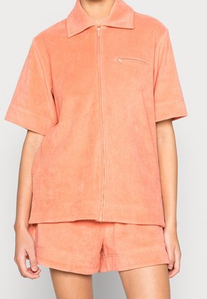 Korte mouwen perzikkleurig terry stoffen shirt met rits en kraag, gecombineerd met bijpassende korte broeken, gedragen door een staand persoon.