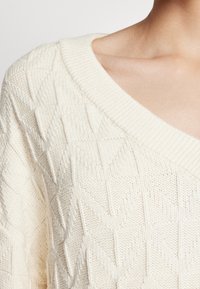 Pull en tricot crème clair avec un col en V, présentant un motif zigzag texturé, un col côtelé et une coupe douce et ample.