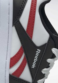 Baskets de sport noires, blanches et rouges avec un matériau en cuir lisse et en maille, rayures latérales proéminentes et lacets. Comprend le logo Reebok sur le côté.